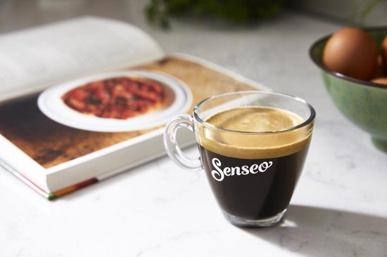 Glas Senseo koffie met schuim op een marmeren aanrecht, met kookboek op de achtergrond.