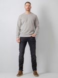 Man met handen in zakken, staand in grijze sweater en zwarte jeans met bruine schoenen.