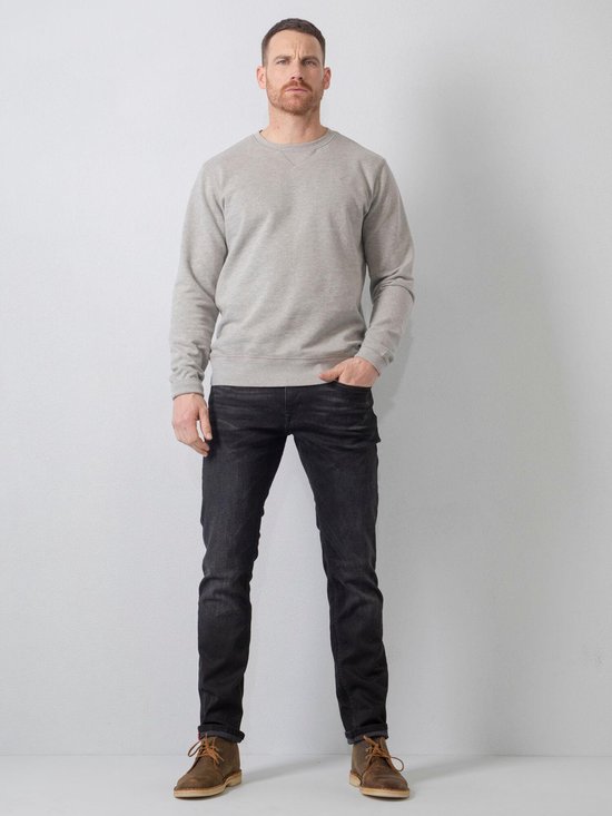 Man met handen in zakken, staand in grijze sweater en zwarte jeans met bruine schoenen.