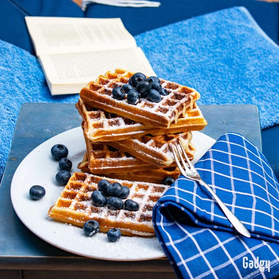 Stapel gouden Brusselse wafels met bosbessen en poedersuiker op een wit bord, met een blauwe servet en vork.