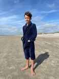 Jongen in een donkerblauwe fleeced badjas met ceintuur, staand op het strand.