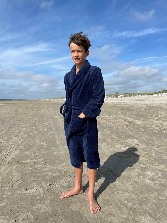 Jongen in een donkerblauwe fleeced badjas met ceintuur, staand op het strand.
