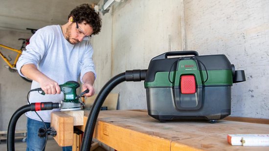 Man met veiligheidsbril schuurt hout, aangesloten op groene en zwarte stofzuiger.