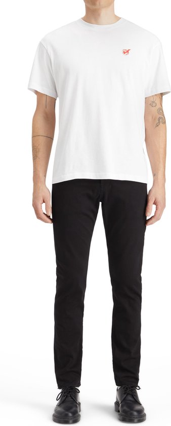 Man in een witte t-shirt met een klein oranje logo en een zwarte jeans.