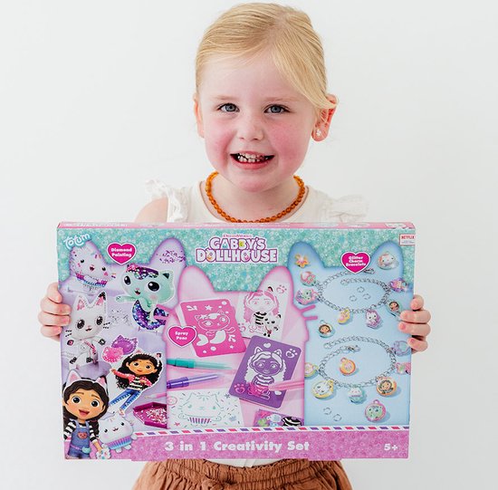 Meisje met blonde staartjes houdt een "Gabby's Poppenhuis 3-in-1 Creativity Set" vast met afbeeldingen van Gabby, Pandy Paws, Cakey Cat en glitter armbandjes.