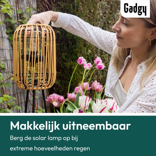 Vrouw zet rotan lantaarn op driepoot in de tuin. "Makkelijk uitneembaar".
