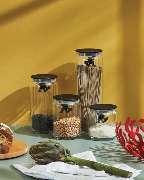 Quatre bocaux en verre avec couvercles noirs. Deux bocaux contiennent des haricots, un contient des spaghettis et un contient du riz blanc. Chaque bocal a une petite figurine noire à l'intérieur du couvercle.
