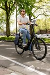Man in blauwe jeans en wit poloshirt rijdt op een zwarte Sparta e-bike met donkerblauwe fietstassen.
