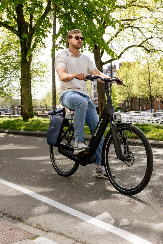 Man in blauwe jeans en wit poloshirt rijdt op een zwarte Sparta e-bike met donkerblauwe fietstassen.