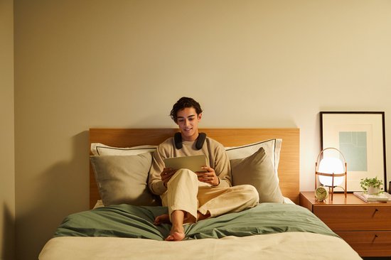 Man met nekband speaker zit op bed een tablet te bekijken.
