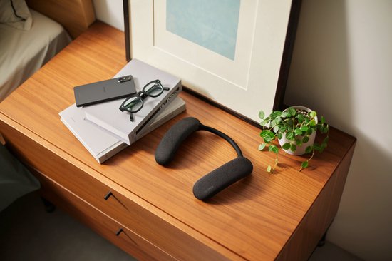 Zwarte nekband speaker op een houten kastje, naast een telefoon, bril en boeken.