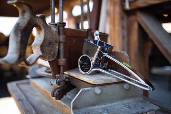Zilverkleurige kaasrasp molen met zwarte greep, naast een oude houten rasp.