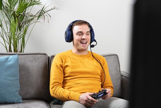 Man met gele trui en blauwe Trust Forze-B gaming headset met camouflagepatroon, spelend op een Playstation.