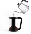 Glazen cafetière met zwarte handgreep, gevuld met gemalen koffie. Water wordt uit een zilverkleurige waterkoker gegoten.