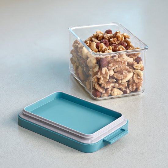 Une boîte de conservation transparente remplie d'un mélange de noix, avec un couvercle turquoise à côté.