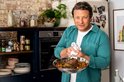 Jamie Oliver, in een blauwe blouse, houdt een braadpan met vlees en groenten omhoog.