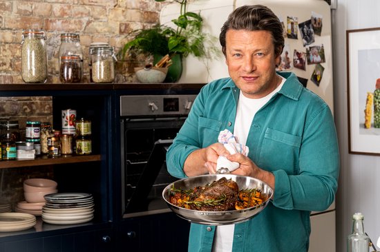 Jamie Oliver, in een blauwe blouse, houdt een braadpan met vlees en groenten omhoog.