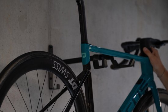 Blauwe racefiets met zwarte velgen hangt aan zwarte fietsmuurbeugel.