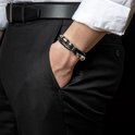 Gevlochten zwarte armband met gouden en zilverkleurige details, gedragen door een man in een wit overhemd.