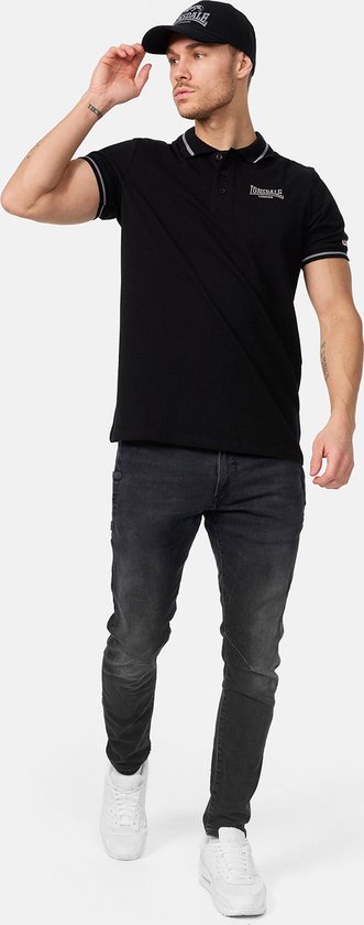 Man draagt zwarte Lonsdale polo met logo, grijze en witte strepen op kraag en mouwen, met zwarte jeans.