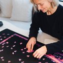Vrouw met blonde bob, glimlachend en puzzelend op een zwarte mat met een paarse en roze puzzel.