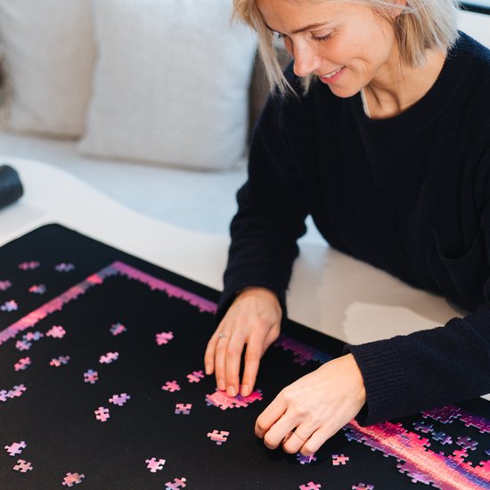 Vrouw met blonde bob, glimlachend en puzzelend op een zwarte mat met een paarse en roze puzzel.