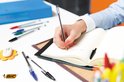Hand schrijft in een open notitieboekje met een BIC Cristal Original pen. Andere pennen en kantoorbenodigdheden op tafel.