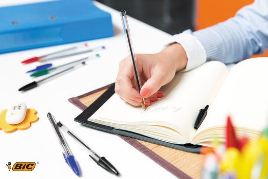 Hand schrijft in een open notitieboekje met een BIC Cristal Original pen. Andere pennen en kantoorbenodigdheden op tafel.