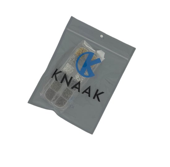 Een gesloten grijze KNAAK-verpakking met een doorzichtig venster waaruit een KNAAK-sieradenset met metalen onderdelen zichtbaar is.