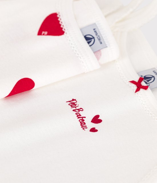 Detailopname van twee witte topjes, één met rode hartjesprint, de ander met 'Petit Bateau' opschrift.