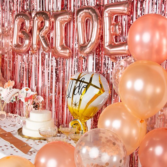 Décoration de table pour fête de despedida de soltera : ballons "BRIDE", diamant "I do", voile de mariée, gâteau et coupes de champagne, avec fond scintillant rose.