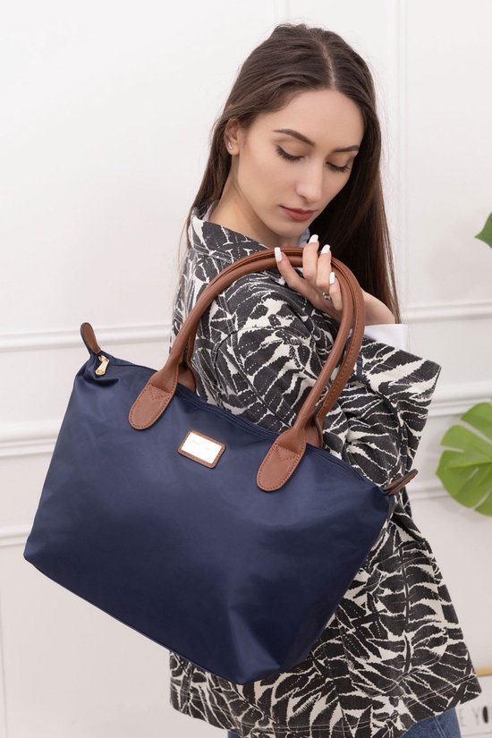 Une femme aux longs cheveux bruns montre un sac en nylon bleu foncé avec des anses en cuir marron.
