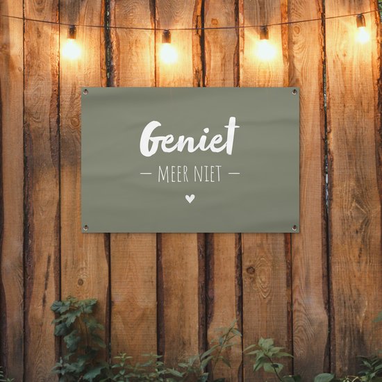 Donkergroene banner met witte tekst 'Geniet - Meer Niet -' en een hartje, hangend aan een houten schutting met lichtslinger.
