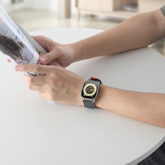 Een persoon draagt een smartwatch met een zwart/grijs sportbandje met oranje accenten, terwijl die een tijdschrift leest.