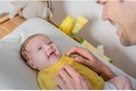 Vrolijke baby lacht op een commode, met een vader die naar hem kijkt. Babyverzorgingsproducten op de achtergrond.