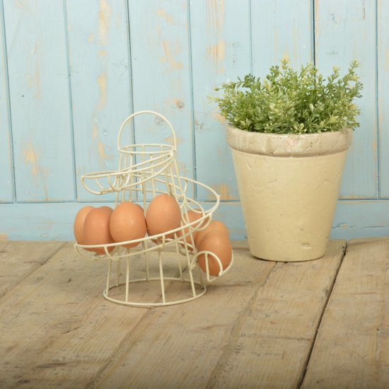 Crèmekleurig metalen spiraal eierrek met 6 bruine eieren, naast een pot met groene plant, op houten ondergrond.