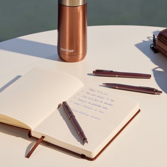 Carnet ouvert avec un stylo bille posé dessus. Deux autres stylos sont à côté. Une bouteille couleur cuivre se trouve derrière le carnet.