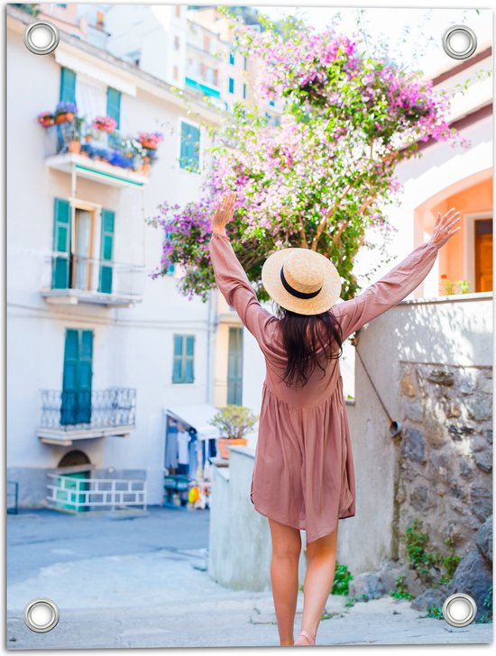 Tuinposter – Juichende Vrouw met Hoed bij Roze Bloemenstruik in Dorpsstraatje - 30x40 cm Foto op Tuinposter (wanddecoratie voor buiten en binnen)