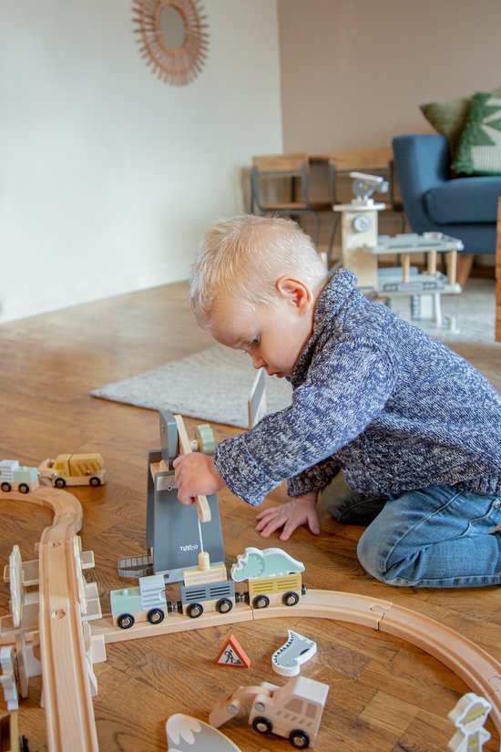 Jongen met blauw vest en spijkerbroek speelt geconcentreerd met houten treinbaan op de vloer.