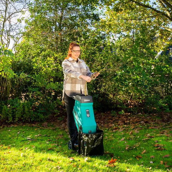 Vrouw met veiligheidsbril in een tuin, gebruikmakend van een groene hakselaar met opvangzak.
