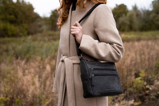 Zwarte leren schoudertas met ritsvakken, gedragen door een vrouw in een beige jas.