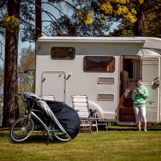 Vrouw in groen vest en witte broek staat buiten naast een camper met een fietshoes voor fietsen ernaast.