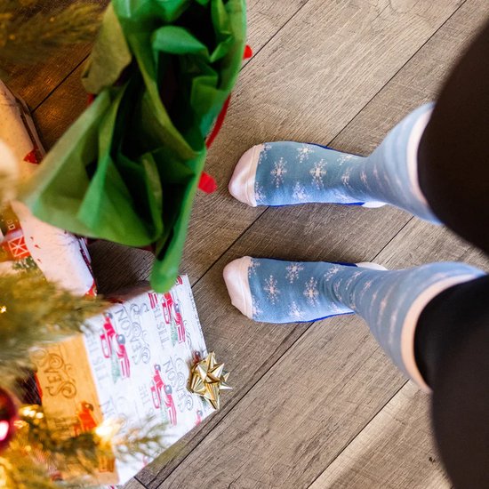 Lichtblauwe kniekousen met witte sneeuwvlokken-opdruk, gedragen door iemand bij een kerstboom en cadeaus.