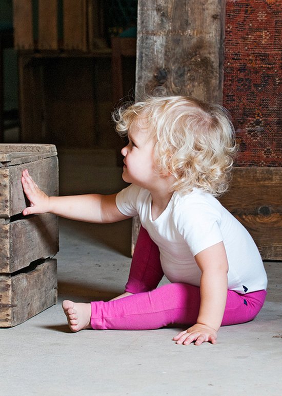 Meisje met blond krullend haar in wit T-shirt en roze legging, zittend naast een houten kist.