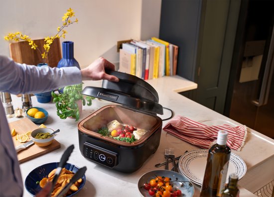 Man opent deksel van zwarte Russell Hobbs multicooker in een keuken. Binnenin stoomt vis met groenten.