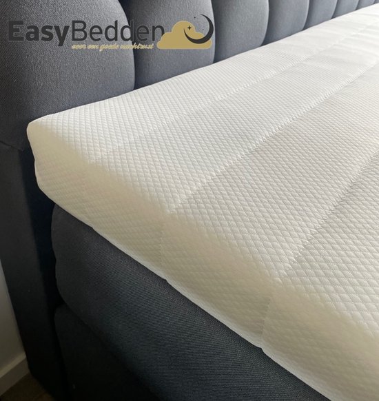 Witte topper met ruitpatroon, op een bed met donkergrijze hoofdbord en matraskast. Easy Bedden logo zichtbaar.