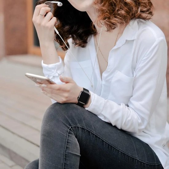 Vrouw met witte blouse, grijze jeans, zittend, met smartphone en zwarte smartwatch.