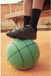 Een kind draagt zwarte schooltennisschoenen met zwarte sokken, zittend op een bankje, met een groene bal onder de voeten.