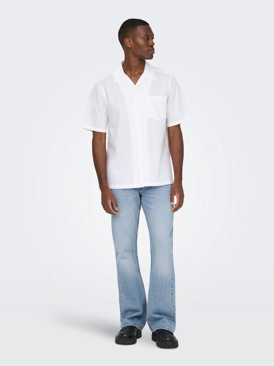 Man met witte korte mouwen blouse en lichtblauwe flared jeans, met de hand in de zak.