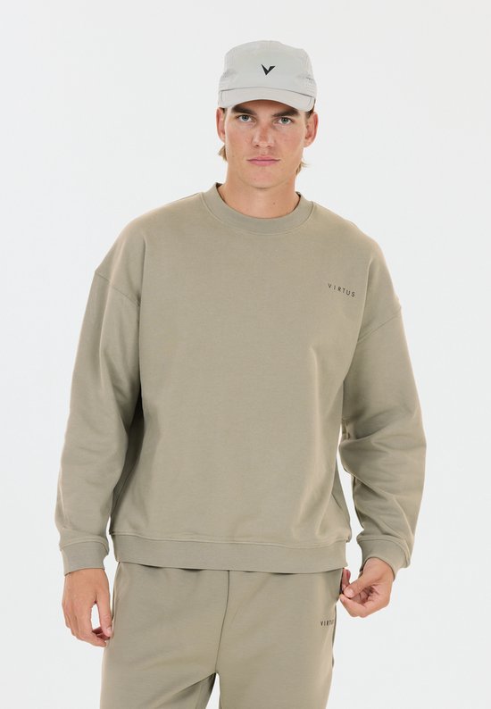 Model in een beige Virtus-set met ronde hals, pet en joggingbroek, kijkend naar de camera tegen een witte achtergrond.